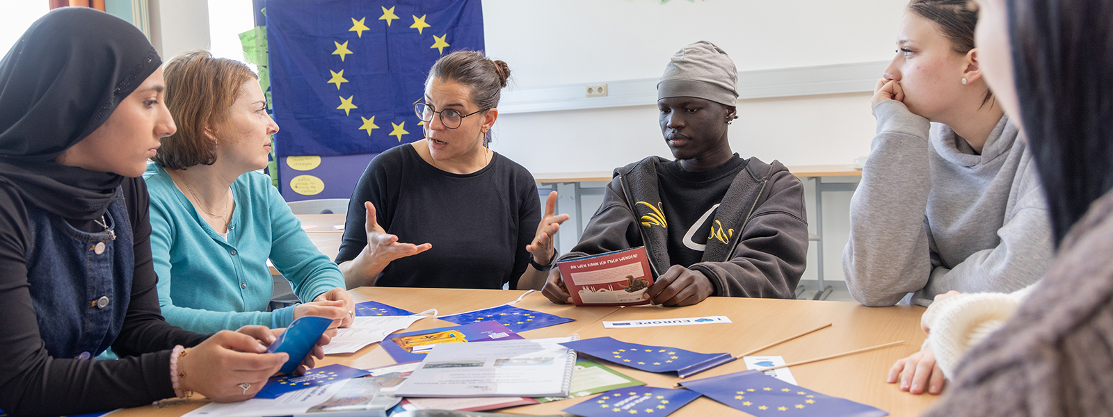 Gespräch zwischen Lehrer/-innen und Schüler/-innen. Im Hintergrund und auf einem Tisch im Vordergrund liegen Europaflaggen
