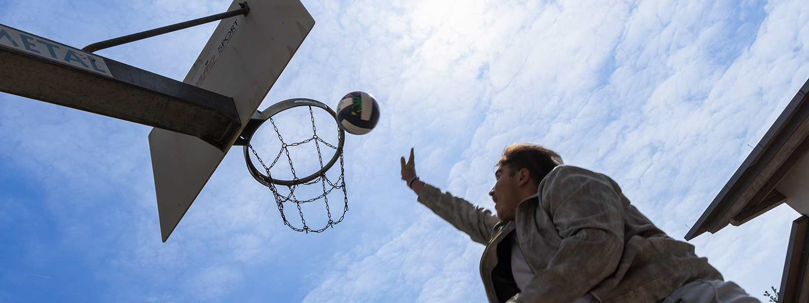 Ein Schüler wirft einen Ball in einen Basketballkorb. Sicht von unten hoch in den blauen Himmel.