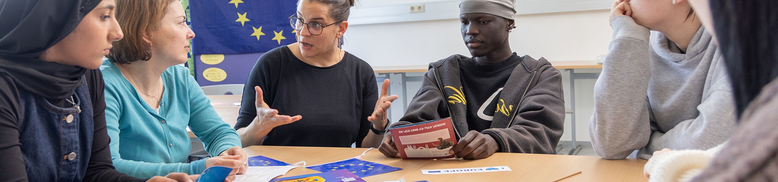 Gespräch zwischen Lehrer/-innen und Schüler/-innen. Im Hintergrund und auf einem Tisch im Vordergrund liegen Europaflaggen
